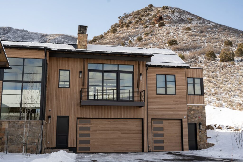 Wide view of a cedar-sided Colorado mountain home with two custom wood garage doors and black-framed windows, installed by American Garage Door & Gate Systems