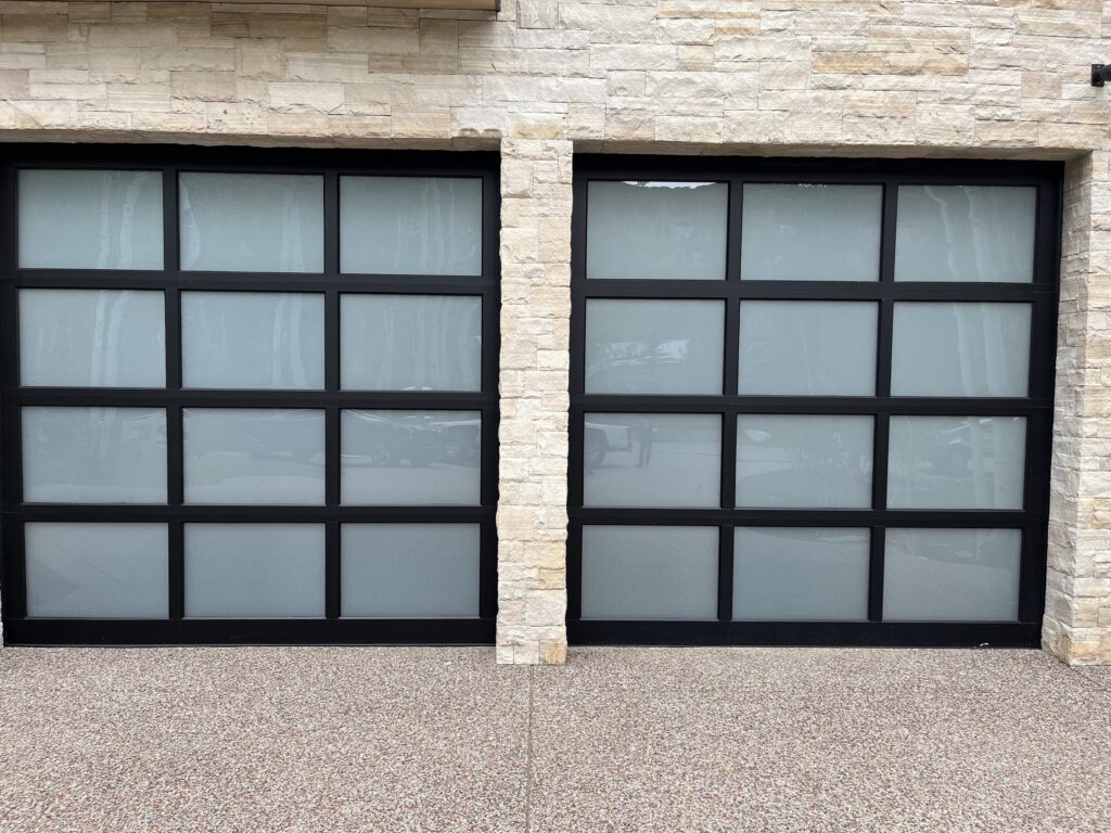 Straight-on view of two frosted glass and black aluminum full-view garage doors set in natural stone, installed by American Garage Door