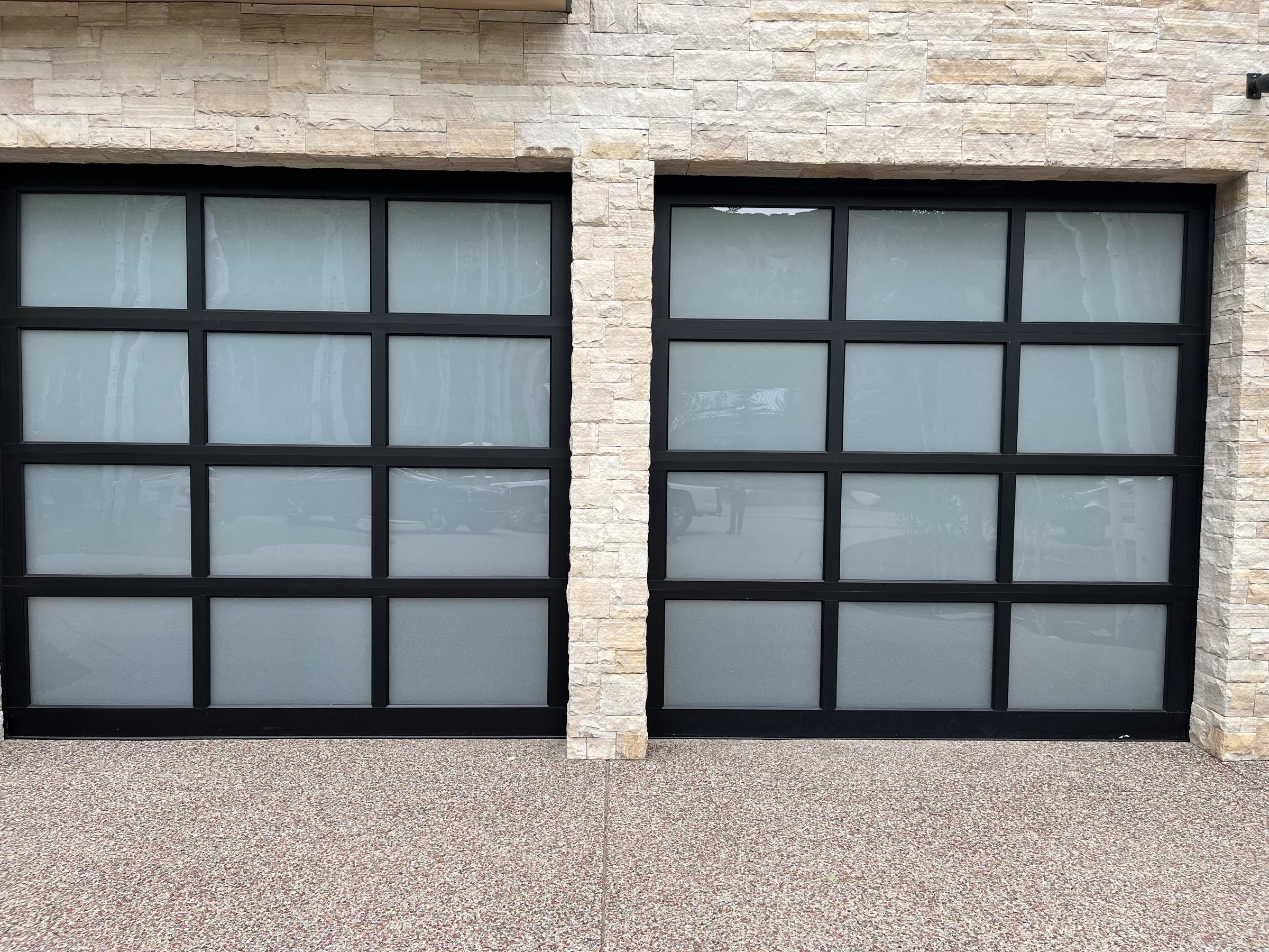 Straight-on view of two frosted glass and black aluminum full-view garage doors set in natural stone, installed by American Garage Door